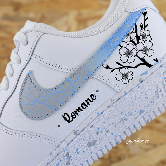 Baskets Nike Air force 1 custom motifs fleurs sakura sur côtés extérieurs avec prénom. Signes Nike peints en dégradé bleu et gris clair avec splash assortis sneakeaze customs
